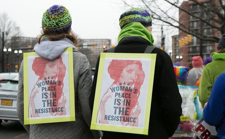 Heron Michelle protesting with Sandwhich board reading A Woman's Place is in the Resistance.