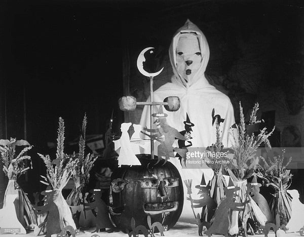 Halloween 1900circa 1905: View of a person, possibly a teacher, wearing a ghost costume behind a lunch table with Halloween decorations in a rural schoolhouse. The display consists of a carved pumpkin, cutouts of witches and black cats, and haystacks. (Photo by Historic Photo Archive/Getty Images)