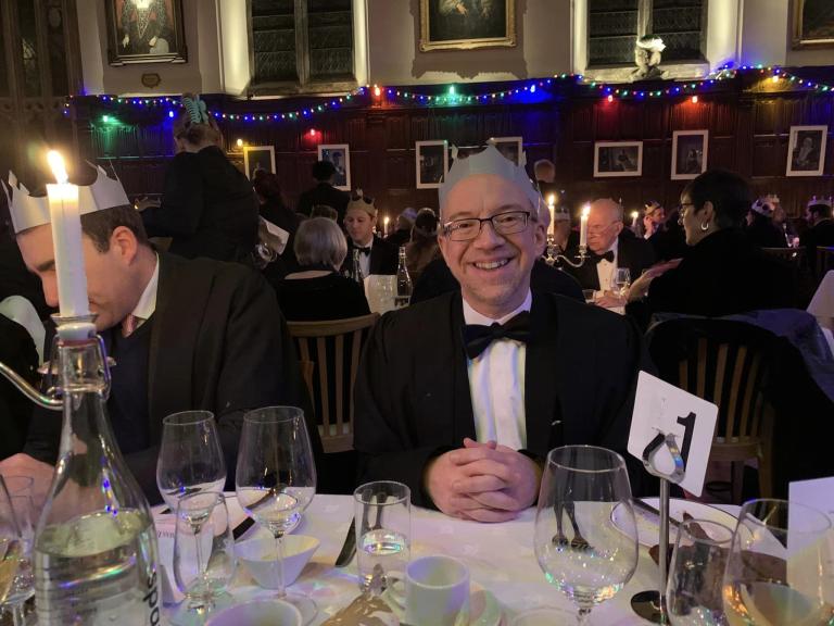 James McGrath sitting in Magdalen College dining hall wearing a paper crown