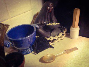 Ancestors altar with dark skinned statuette, blue bowl, bag of graveyard soil, list of honored dead, a cross, and a spoon made of shell.