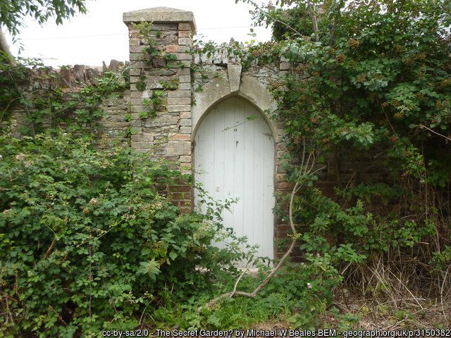 An image of a white wooden door in a grey stone wall surrounded by greenery.