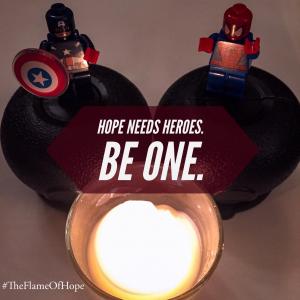 a candle lit in front of two lego superhero minifigs with the words Hope Needs Heroes. Be one.