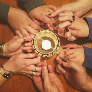 A group of people holding hands around a lit candle. Their hands make a circle around the candle.