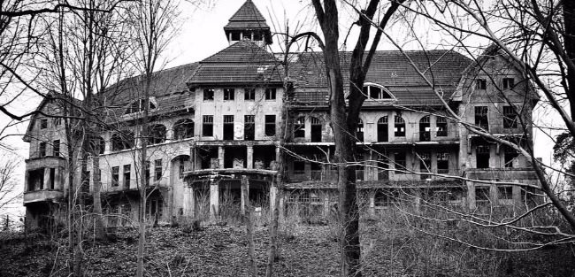 Black and white photograph of a dilapidated 3 story sprawling wooden mansion. The lifeless trees and empty windows only make it creepier. 