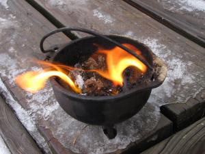 A small cauldron sits on the corner of an icy porch. A tiny fire is burning within it. 
