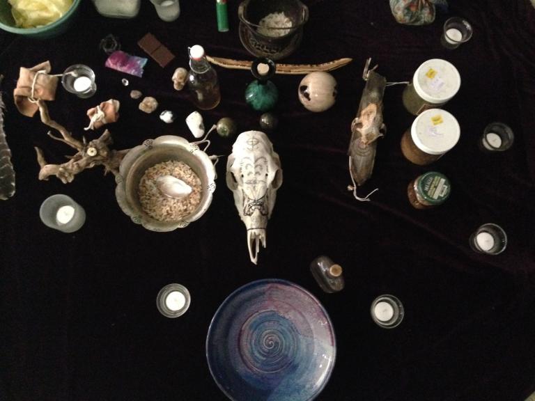 An altar strewn with skulls, candles, and offerings. Jars and stones share room with small spirit bundles. 