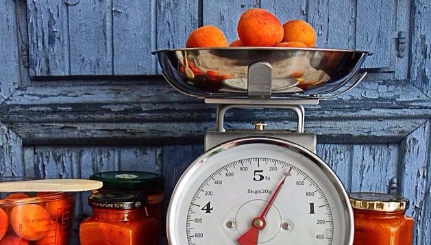 A worn painted blue door stands behind a harvest of peaches. A metal scale is weighing some while others are already canned or turned into jelly. 