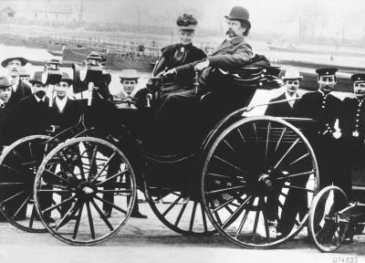 Bertha and Carl Benz sitting in one of their horseless carriages, surrounded by various onlookers.