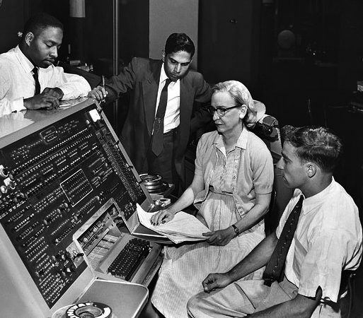 Grace Hopper sits at a large 50s style computer along with three men in suits. She is holding a sheaf of papers. 