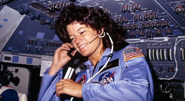 A young and smiling woman is talking into her headset wearing a blue NASA jumpsuit. She is in a small cockpit area with many switches and wires.