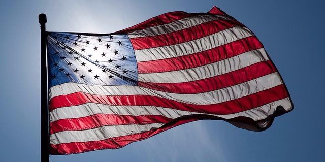 A United States flag blowing in the wind backlit by the sun in a blue sky.