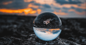A clear marble sits on the soil and inverts the image of the sun that is setting in the background.