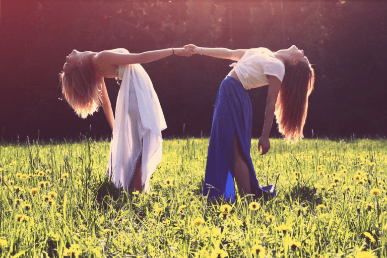 Two women holding hands and leaning away so that it is only their held hands that keep them upright. They are standing in a sunny bright field.