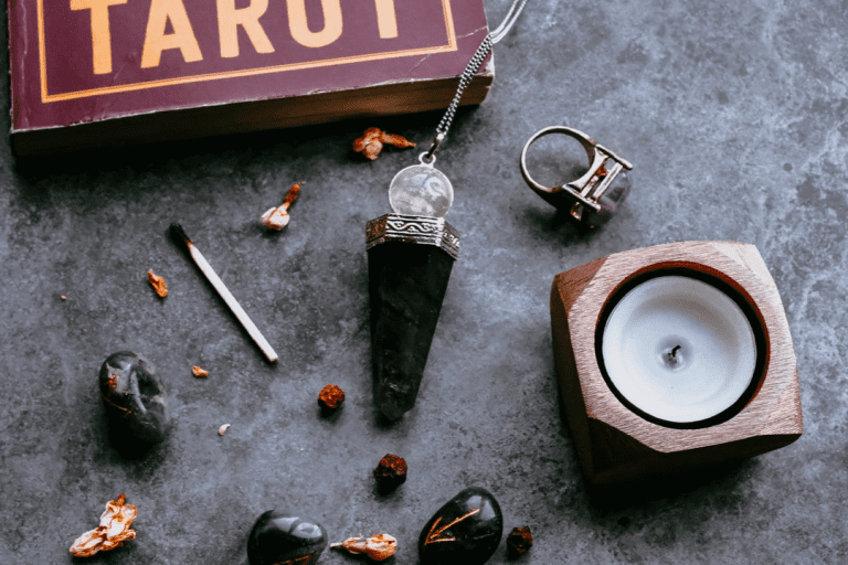 A photo of various accoutrements of ritual: a tarot book, pendulum, burnt out match and candle, runes, and an amethyst ring.