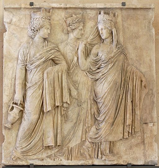 A pale stone bas relief of three goddesses in finely detailed robes with crowns. One carries a pitcher.