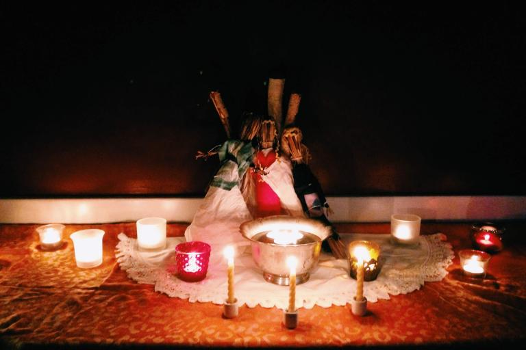 Many candles are lit around a silver bowl with more candles floating in it. Three rustic dolls are made of grass and linen.