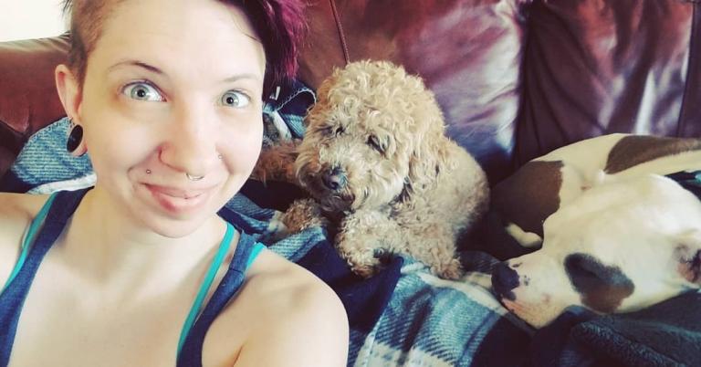 myself, a femme with red-purple hair, smiling into the camera while a brown goldendoodle and a pit bull cuddle behind me