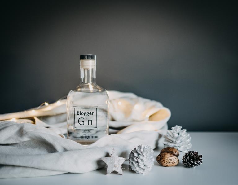 A bottle labeled "Blogger Gin" draped in a white cloth with frosted pine cones beside it.