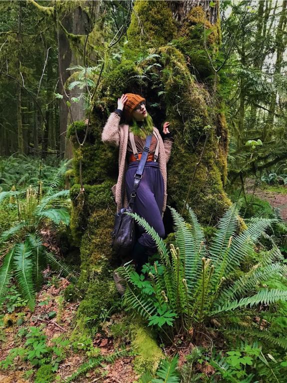 a pale witch in bohemian clothing standing n the hollow of a mossy green tree