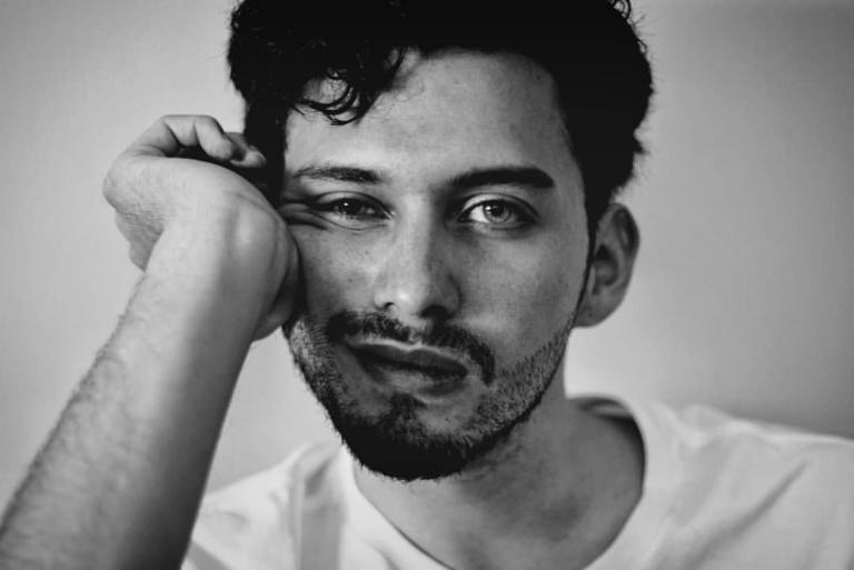 a black and white photo of a scruffy male witch/theurgist bracing his cheek on his hand