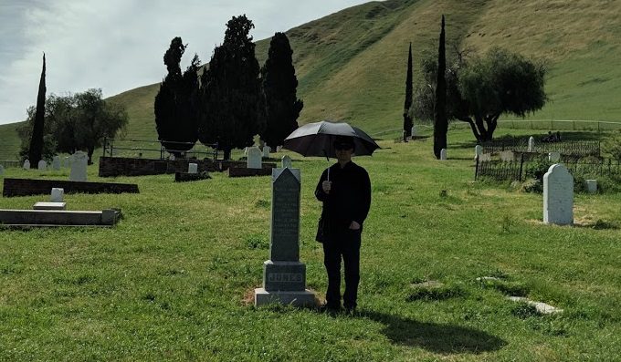 A man stands next to a grave with an umbrella protecting him from the glaring sun.