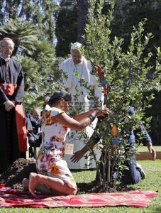 Pope Francis looks on while two Amazon indigenous persons ceremonially add soil and water to newly planted tree.