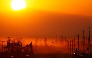 A field of oil rigs backlit by the orange glow of sunset and polluted sky.