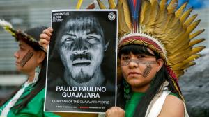 Woman with painted face wearing feathered headdress holds poster of Paulo Paulino Guajajara.