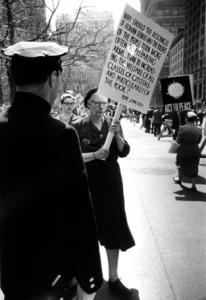 A police officer faces civil defense protesters and Dorothy Day holds sign, with a statement of Pope John XXIII decrying money and talent spent on war.